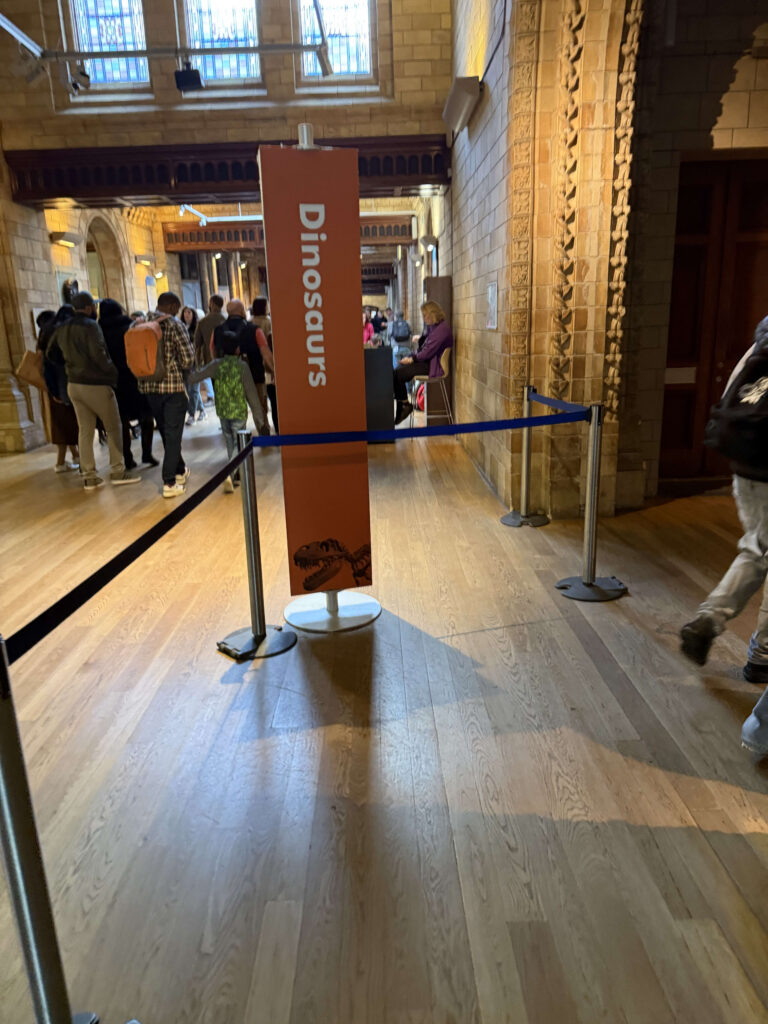 A vertical orange sign reading 'Dinosaurs' standing in a grand museum hallway with a queue of visitors behind blue stanchions.