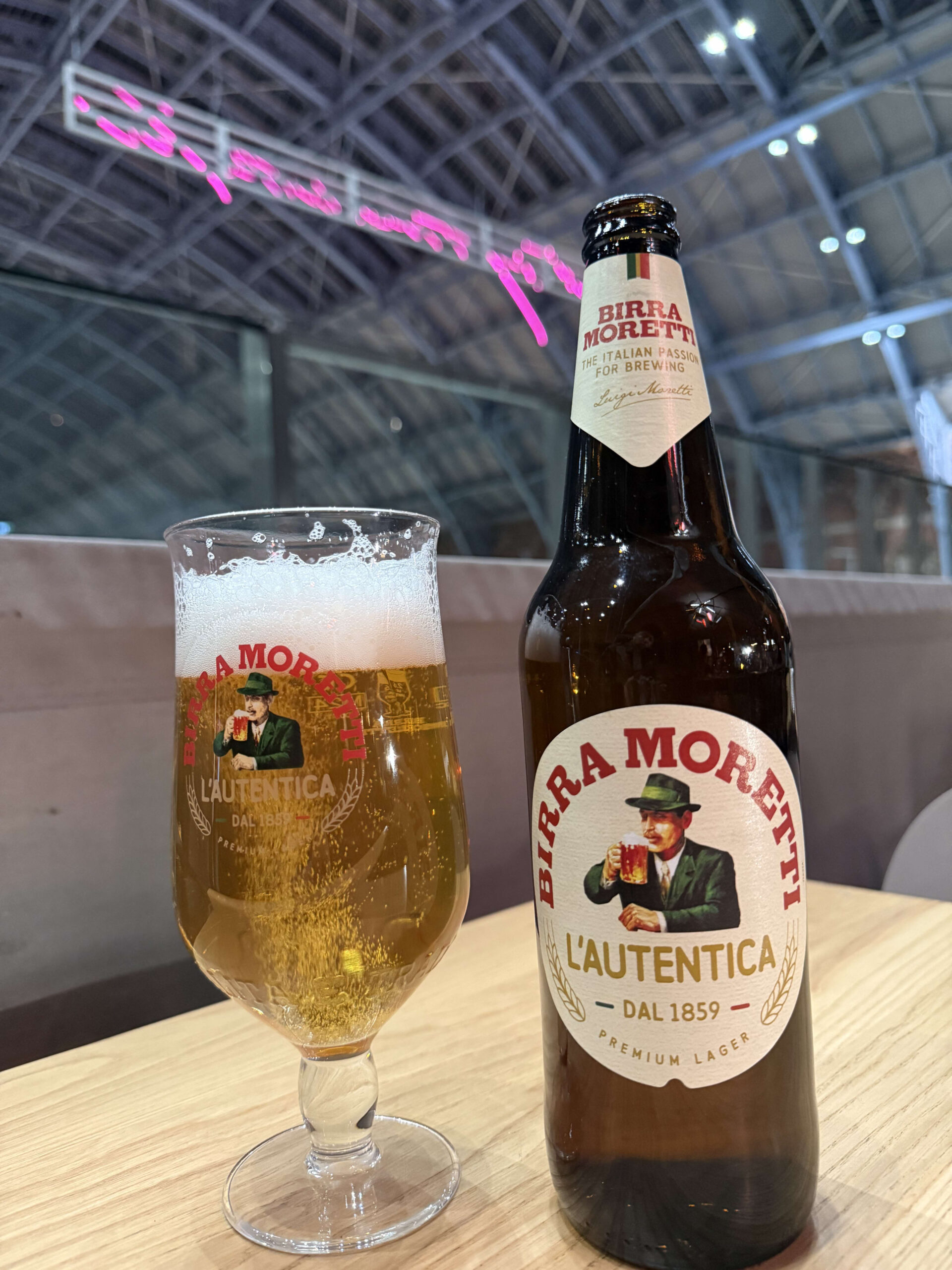 高い天井とピンクのネオンサインを背景に、木製のテーブルに置かれたモレッティビールのボトルとビールが注がれたグラス