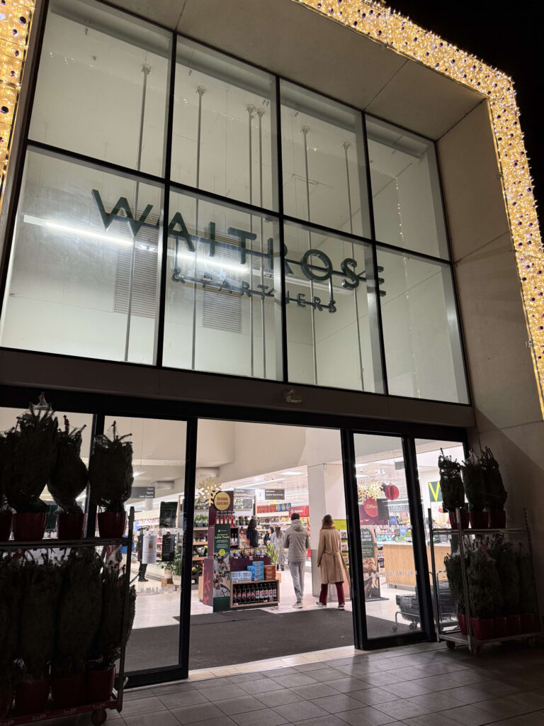 夜のライトアップされたWaitrose & Partners ザ・ブランズウィック・センター店の外観。大きなガラス窓とロゴ、入口付近に並ぶ販売用のクリスマスツリー。