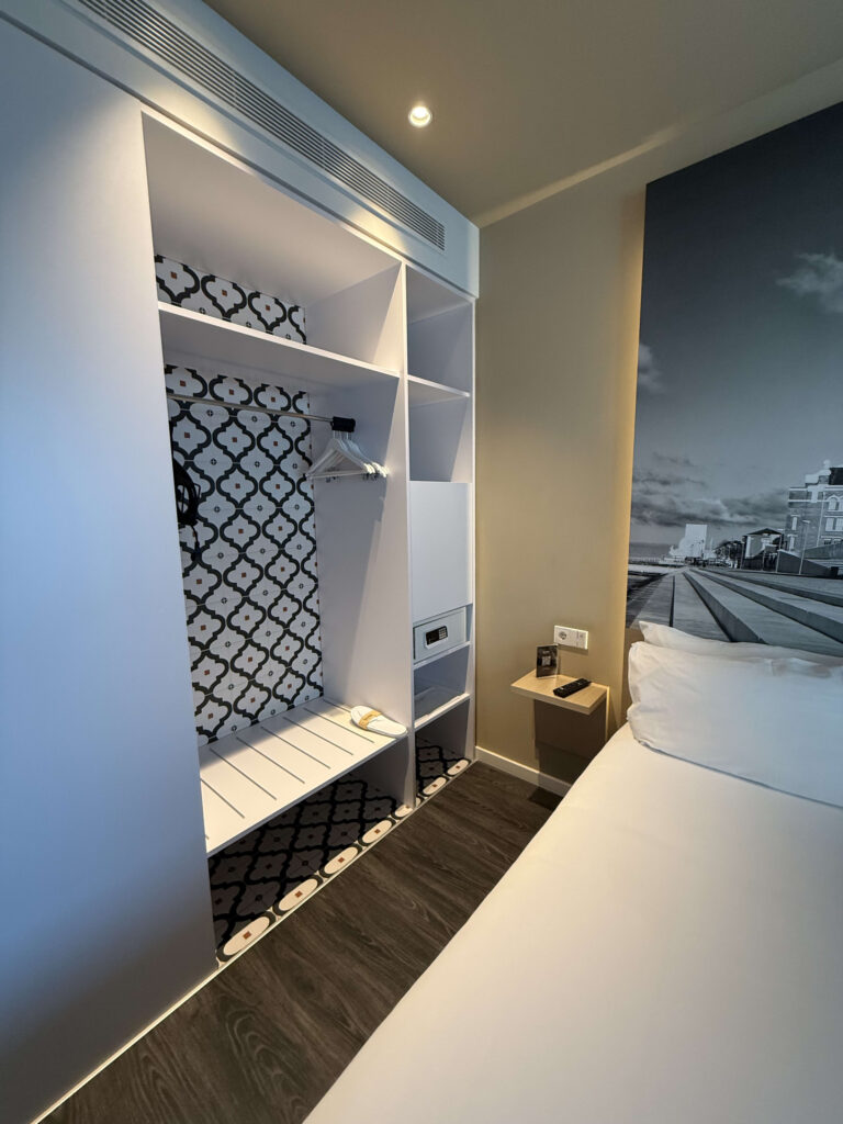 White open wardrobe with geometric pattern backing and hangers next to a hotel bed