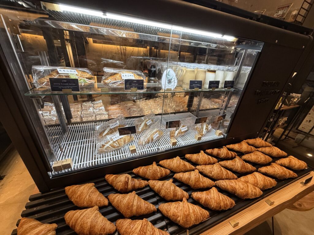 A bakery display case filled with sandwiches and desserts, with rows of fresh croissants in the foreground