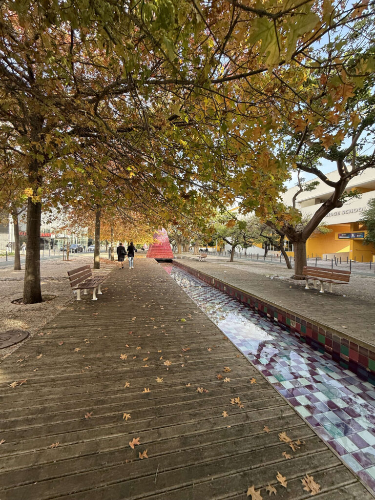 Wooden boardwalk lined with autumn trees and a tiled water channel along Alameda dos Oceanos near MEO Arena