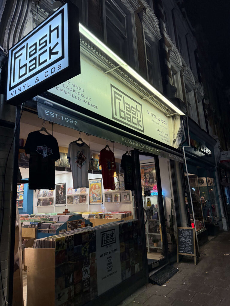 Exterior of Flashback Records store at night, featuring a bright illuminated sign, hanging t-shirts in the window, and racks of vinyl records visible inside.