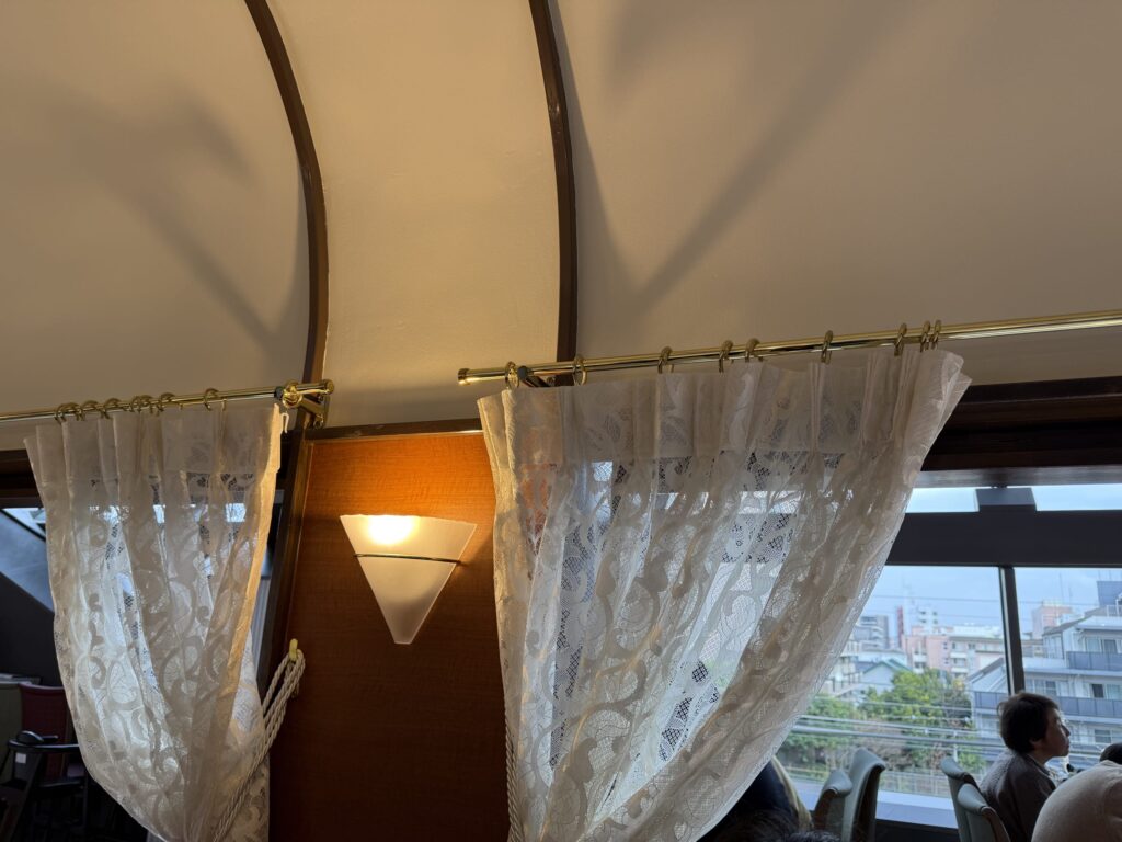 Interior of a restaurant designed like a vintage train dining car, featuring lace curtains, brass luggage racks, and warm lighting.