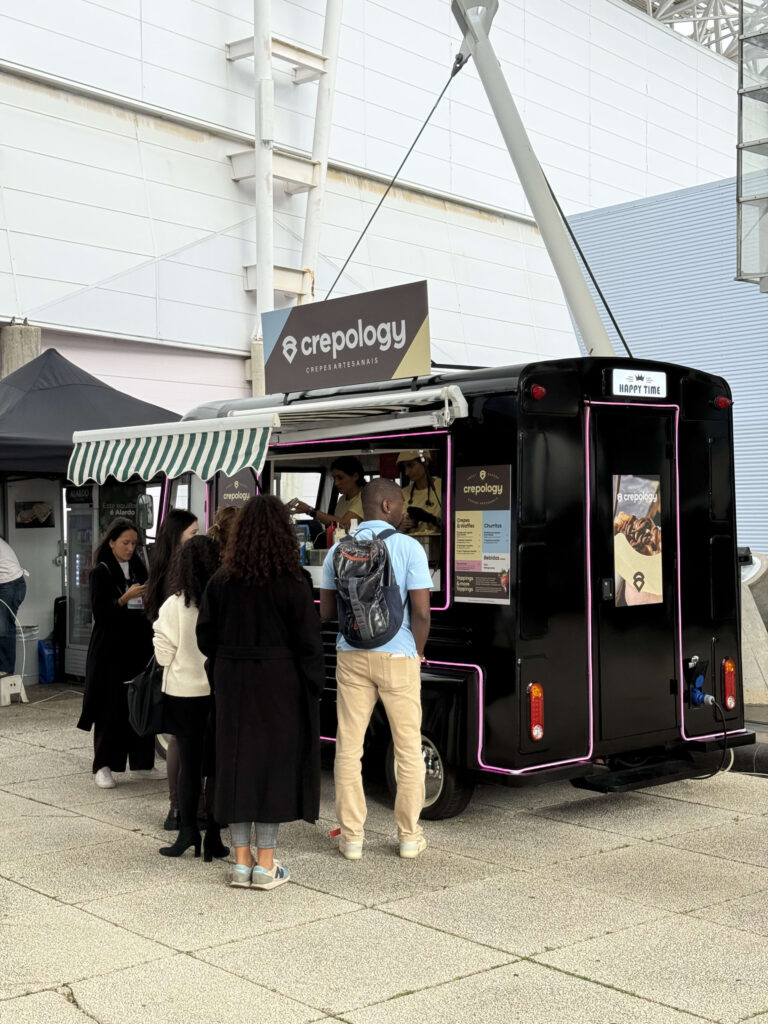People standing in line at a black food truck named 'crepology' with pink neon lights parked outdoors.