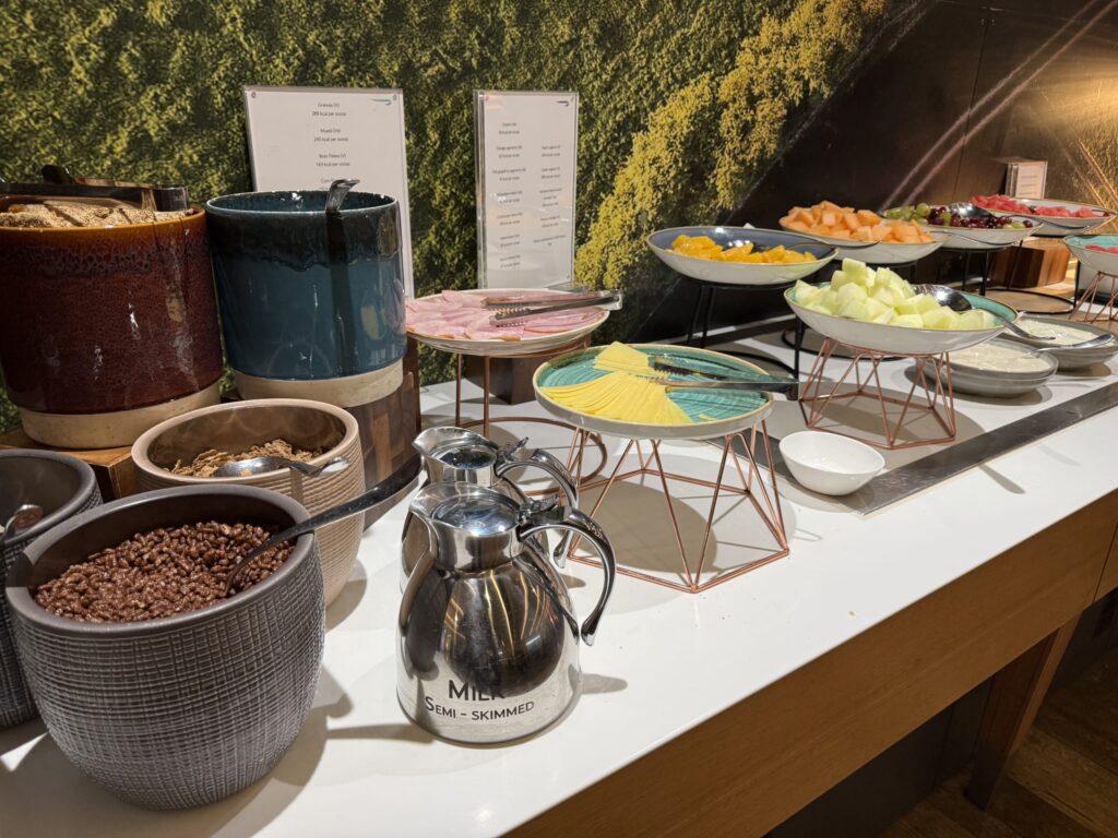 Breakfast buffet counter featuring bowls of cereal, sliced ham, cheese, cut fruits, and a semi-skimmed milk jug