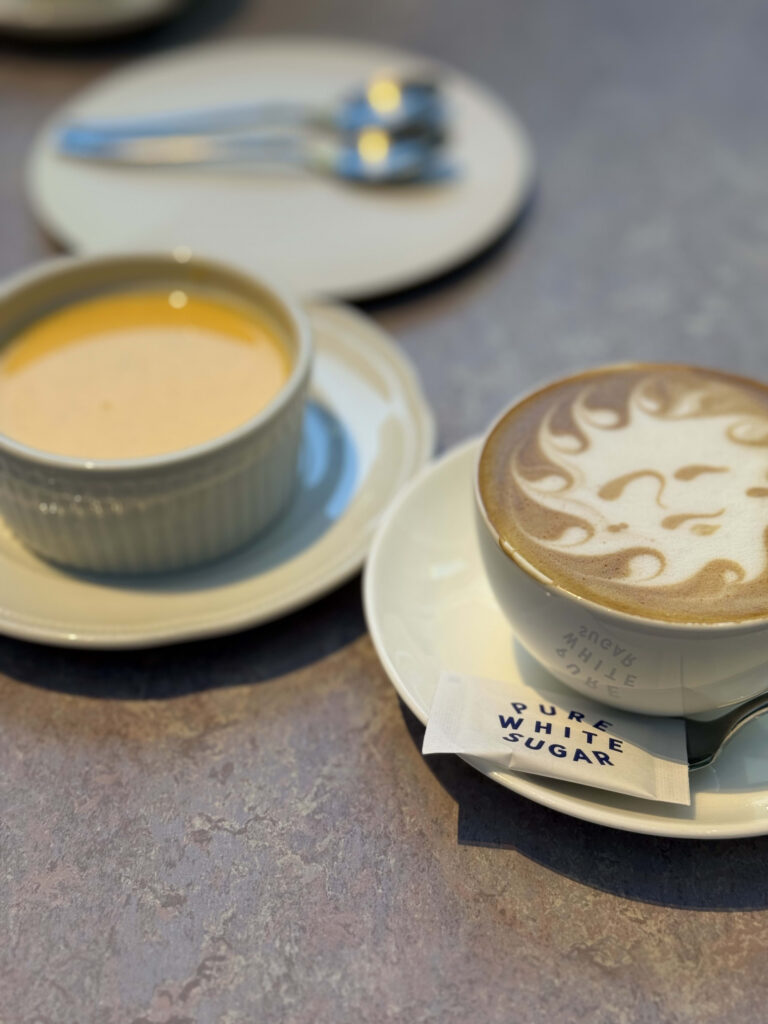 Cup of Koji latte featuring sun-face latte art with a yellow soup or dessert in the blurred background