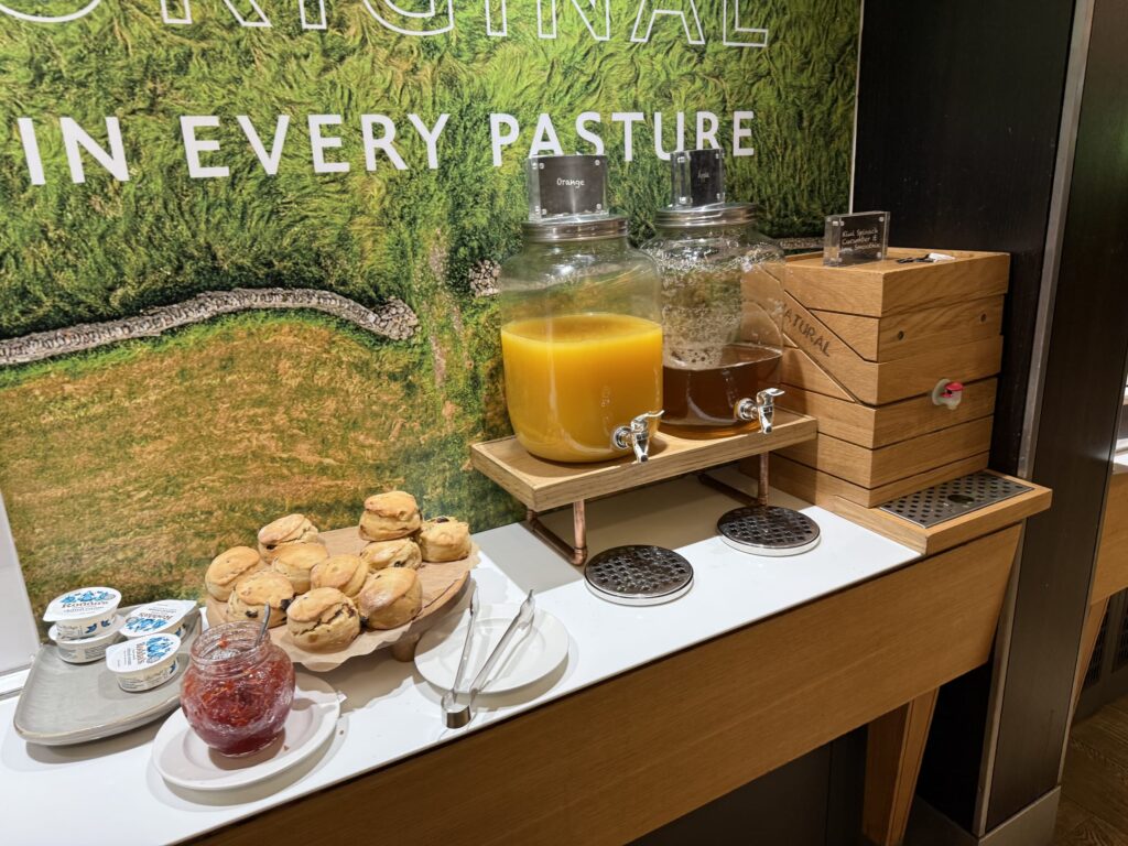 ホテルの朝食ブッフェカウンターに並ぶ、山盛りのスコーン、Rodda'sのクロテッドクリーム、ジャム、そしてオレンジジュースなどのドリンクサーバー