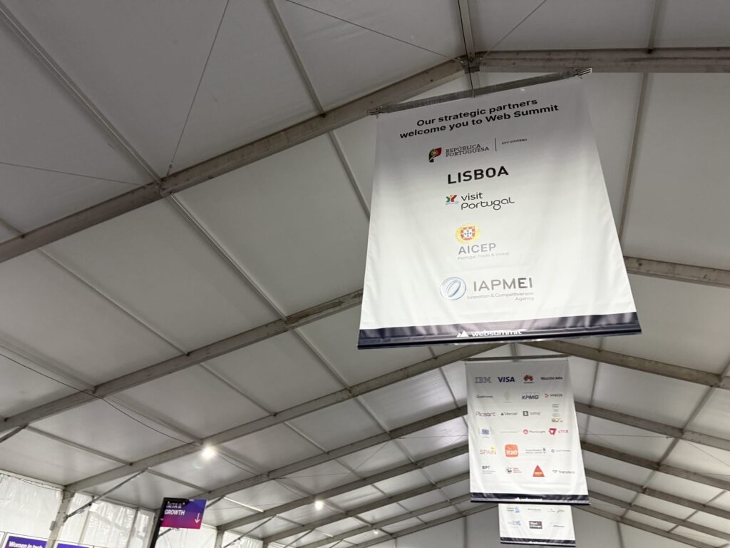Large white banners hanging from the ceiling at the Web Summit venue, featuring logos of strategic partners such as the Portuguese government and the city of Lisbon
