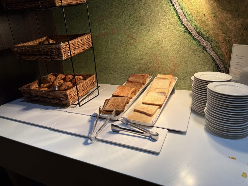 Bread station at a hotel breakfast buffet featuring baskets of croissants and trays of toasted bread next to stacked plates.
