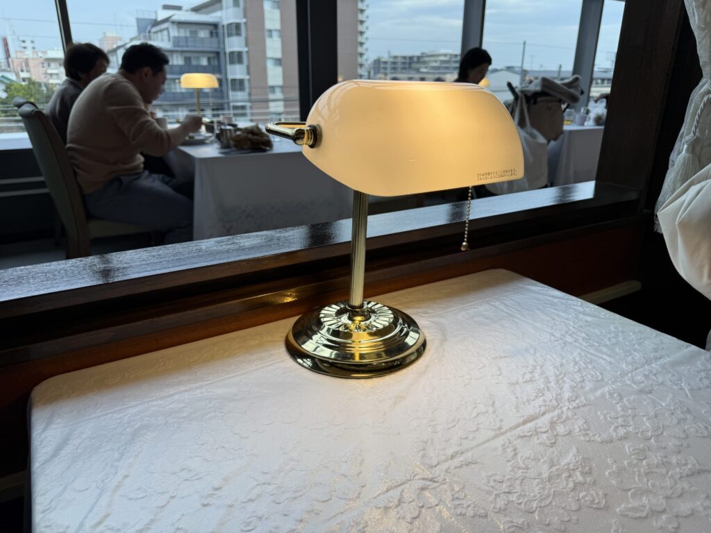 白いレースのテーブルクロスの上に置かれた、温かい光を放つ食堂車風のレトロな真鍮製卓上ランプ