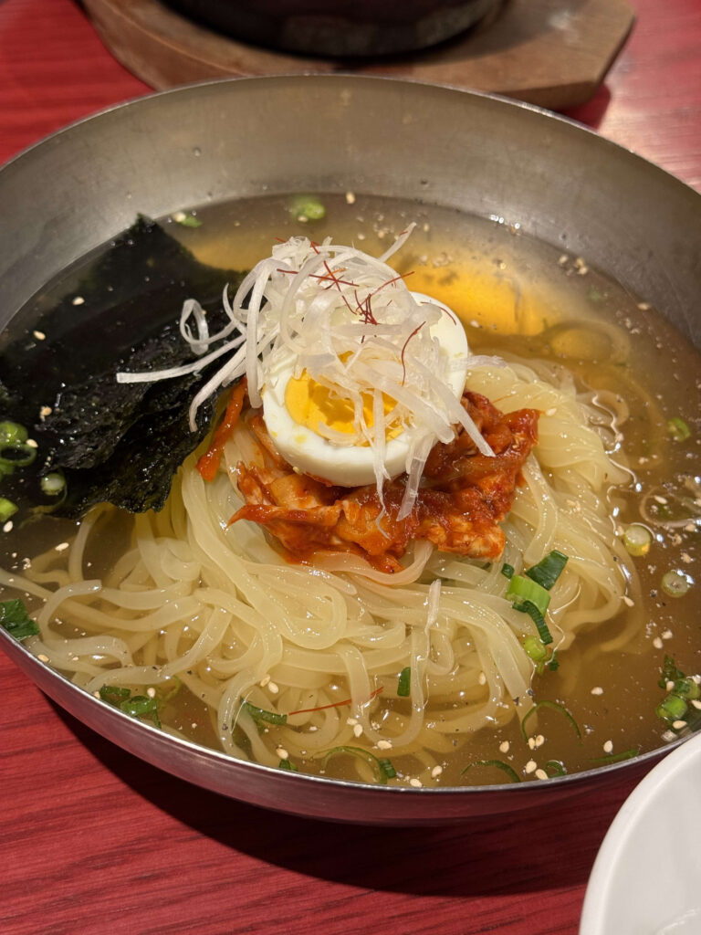 Cold noodles in a stainless steel bowl topped with a hard-boiled egg, kimchi, seaweed, and shredded white leeks in a clear broth.