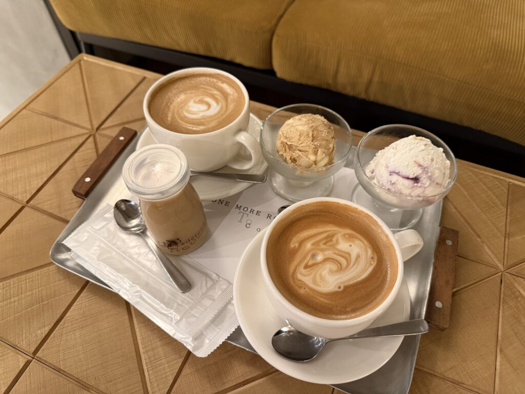 A silver tray on a geometric wooden table holding two cups of coffee with latte art, two scoops of ice cream in glass bowls, and a jar of coffee pudding.