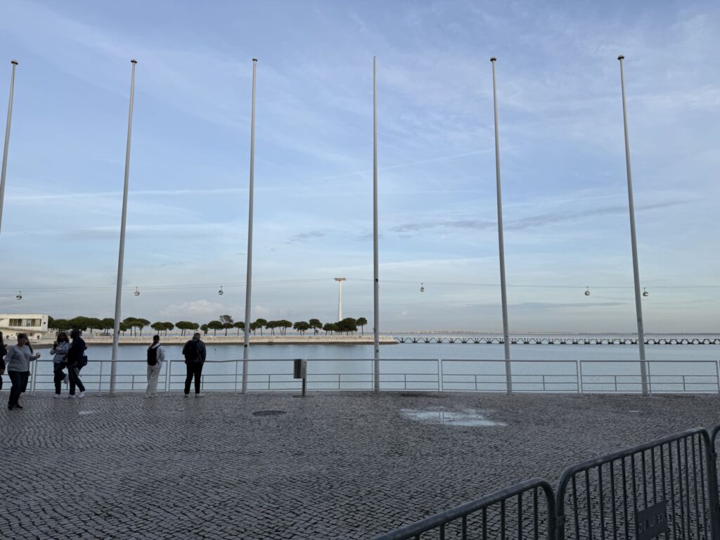 A row of tall white flagpoles on a cobblestone plaza along the Tagus River with cable cars visible in the distance