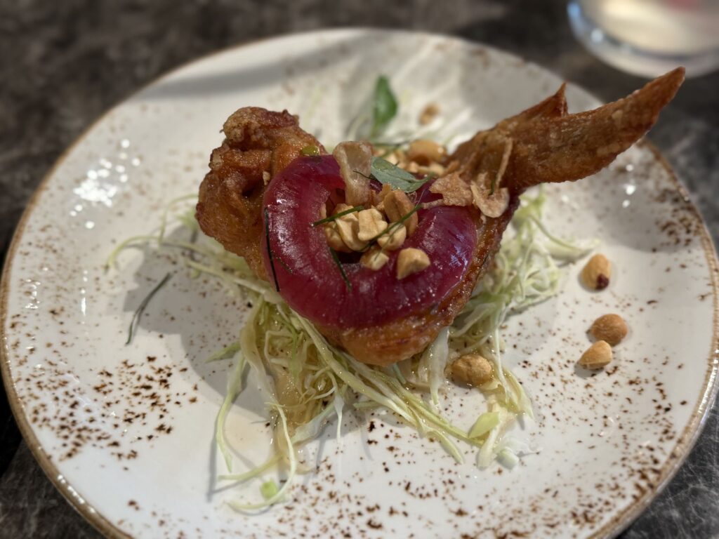 Fried chicken wing appetizer topped with a red onion ring, crushed nuts, and herbs on a speckled plate