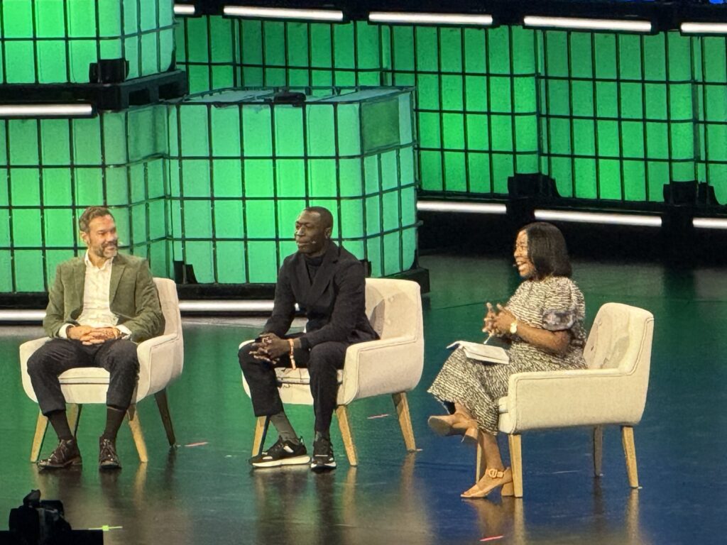 TikToker Khaby Lame sitting on a white chair between two other speakers on a stage with a green illuminated background