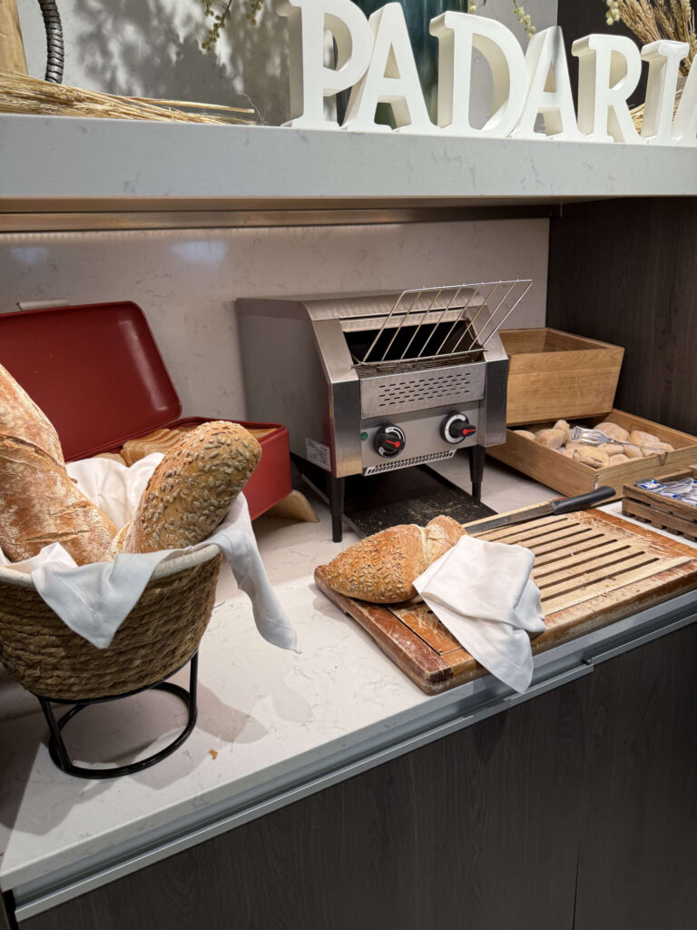 A bakery corner featuring a basket of baguettes and seeded bread, a stainless steel toaster, and a wooden cutting board under a PADARIA sign