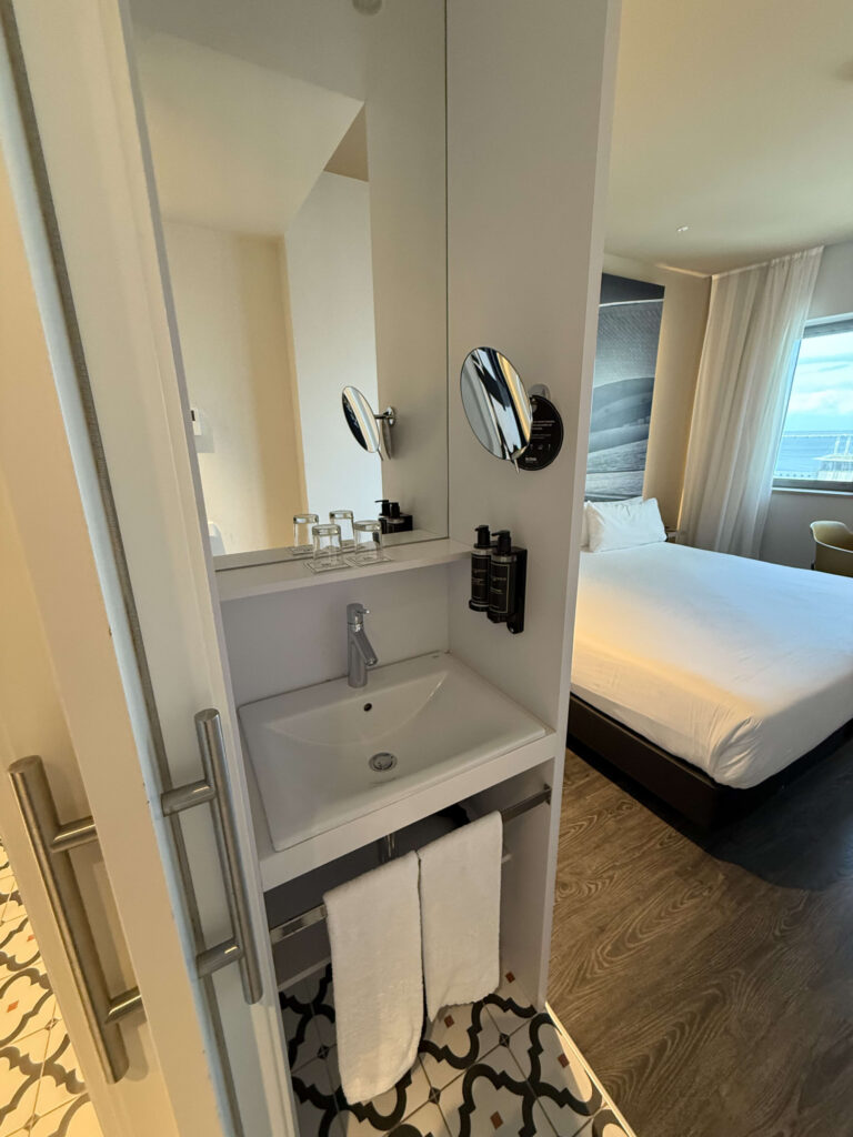 A white hotel bathroom sink with a mirror, amenities, and a view of the bedroom in the background