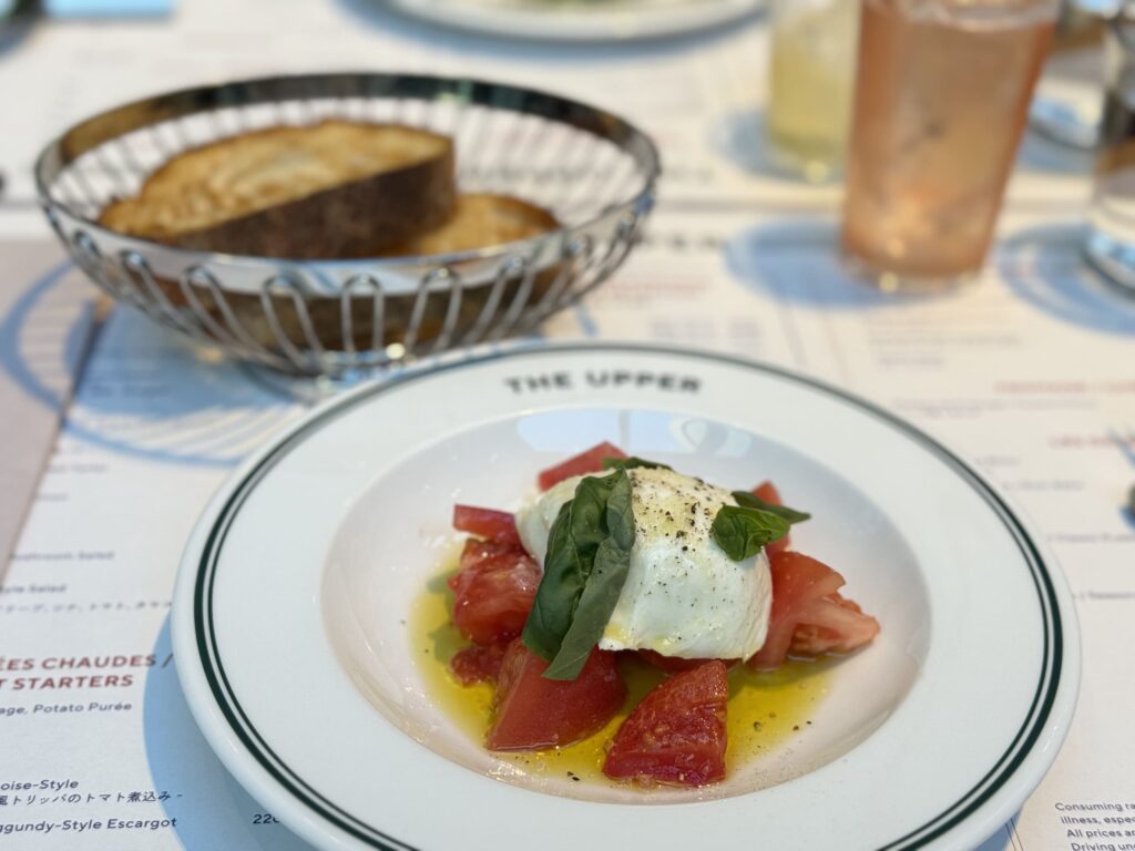 「THE UPPER」のロゴが入った白い皿に盛られた、ブッラータチーズとトマトのカプレーゼと背景のパン