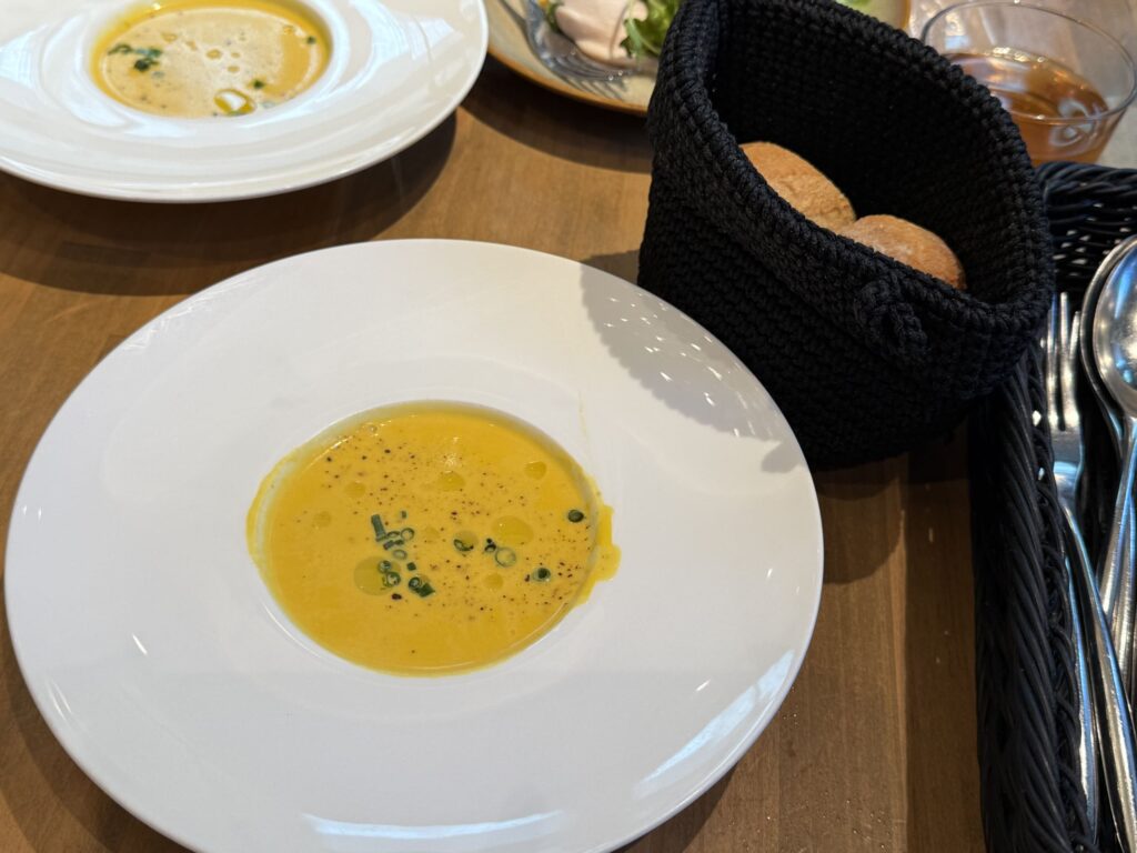 木製テーブル上の白い皿に盛られたカボチャのポタージュと黒い籠に入ったパン