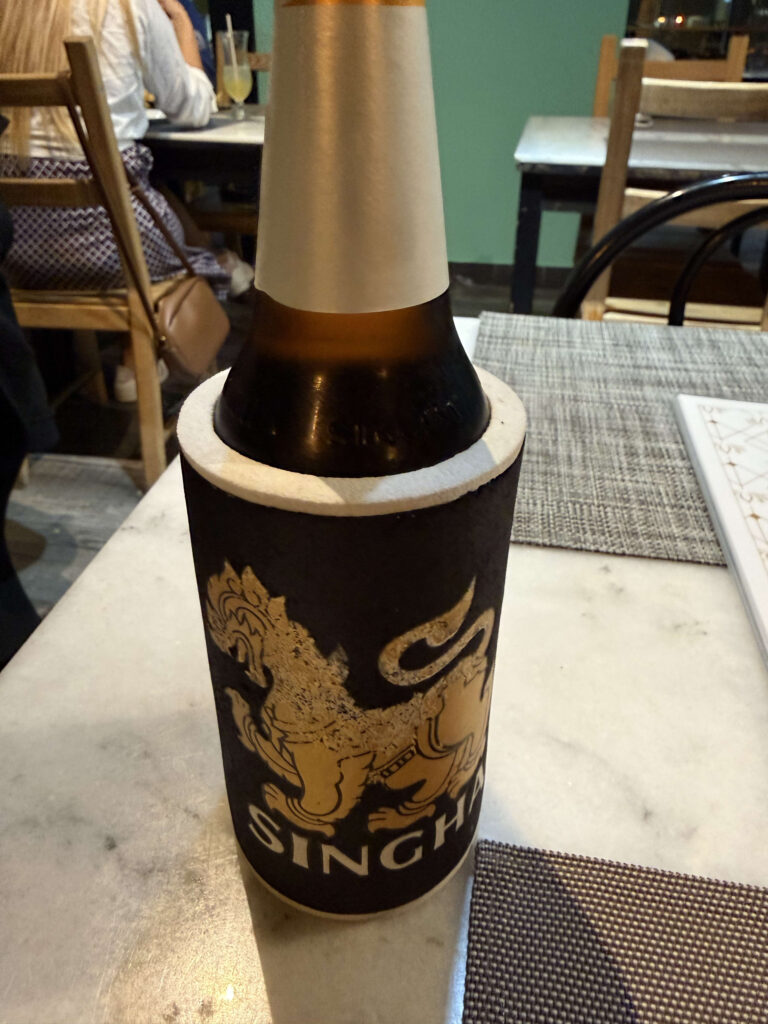 A bottle of Singha beer inside a black koozie holder sitting on a white marble table in a restaurant