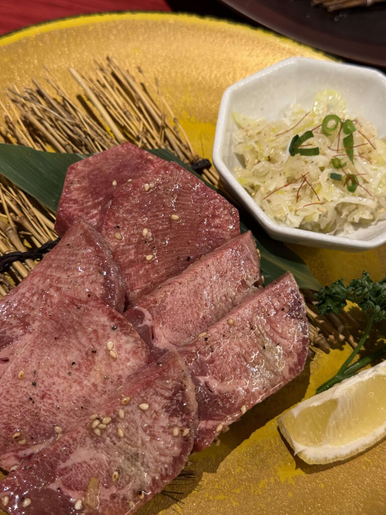 Thick-cut raw beef tongue slices seasoned with sesame seeds on a golden plate with a side of chopped green onion salt at Yakiniku Toraji