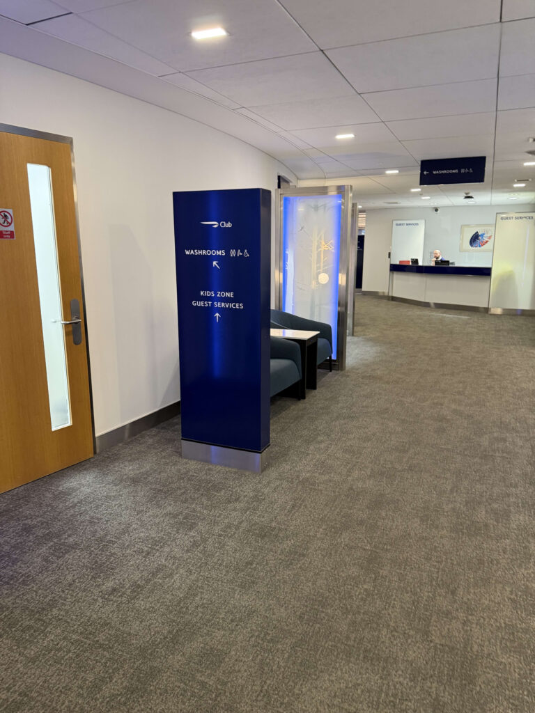 Blue directional signage in the British Airways Club lounge indicating directions to Washrooms, Kids Zone, and Guest Services
