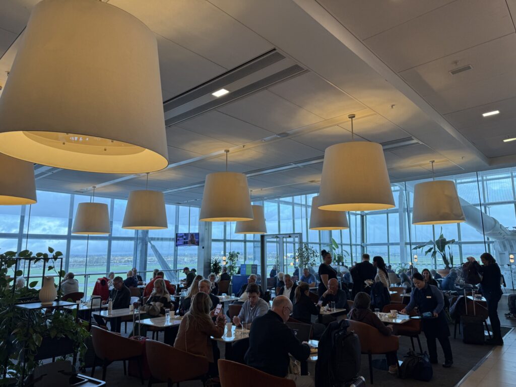 Crowded airport lounge with high ceilings, large windows, and oversized pendant lights