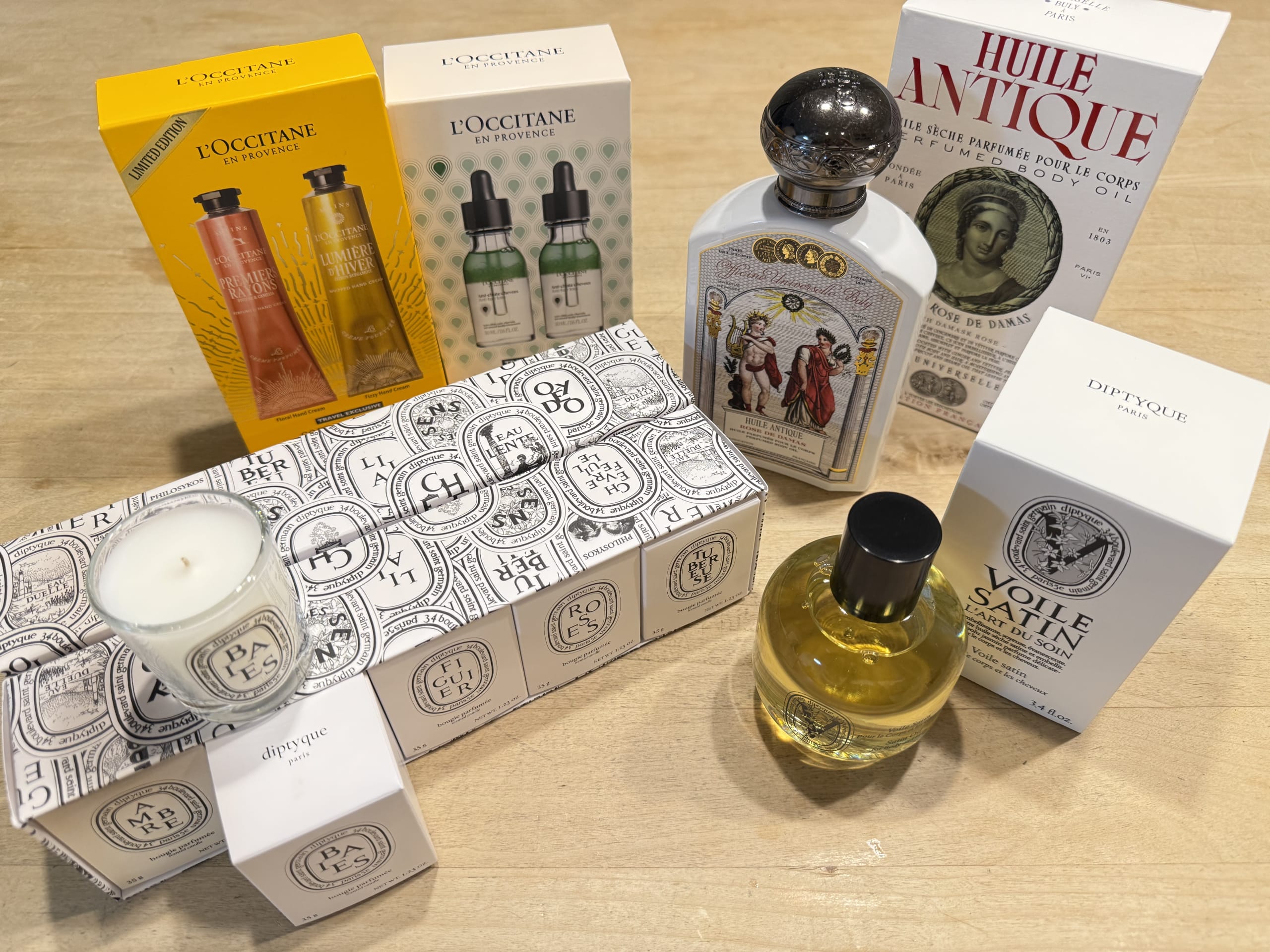 A collection of L'Occitane hand creams and serum, Buly 1803 body oil, and Diptyque candles and satin oil arranged on a wooden table, purchased from JAL in-flight sales