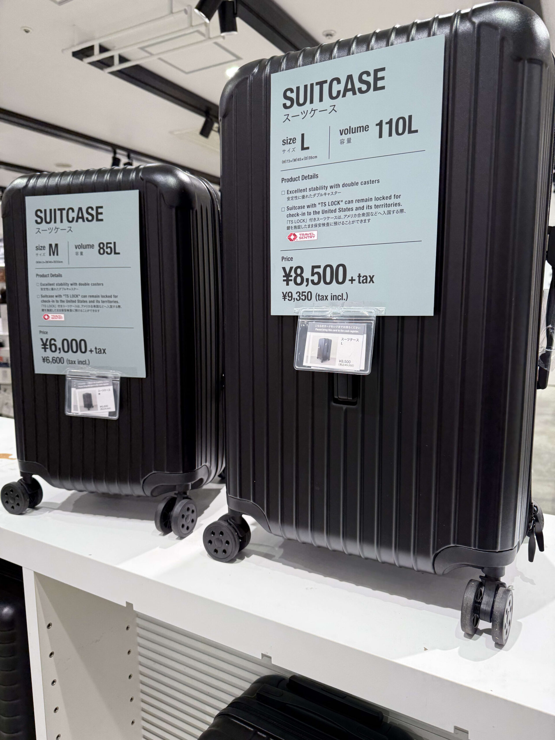 Display of two black hard-shell suitcases, size M (85L) and size L (110L), with price tags attached.