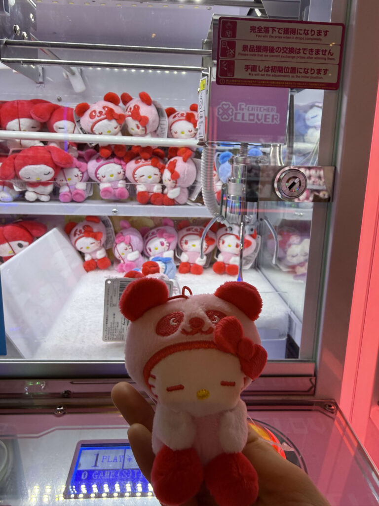 クレーンゲーム機の前で、ピンク色のパンダの着ぐるみを着たハローキティのぬいぐるみを手に持っている様子