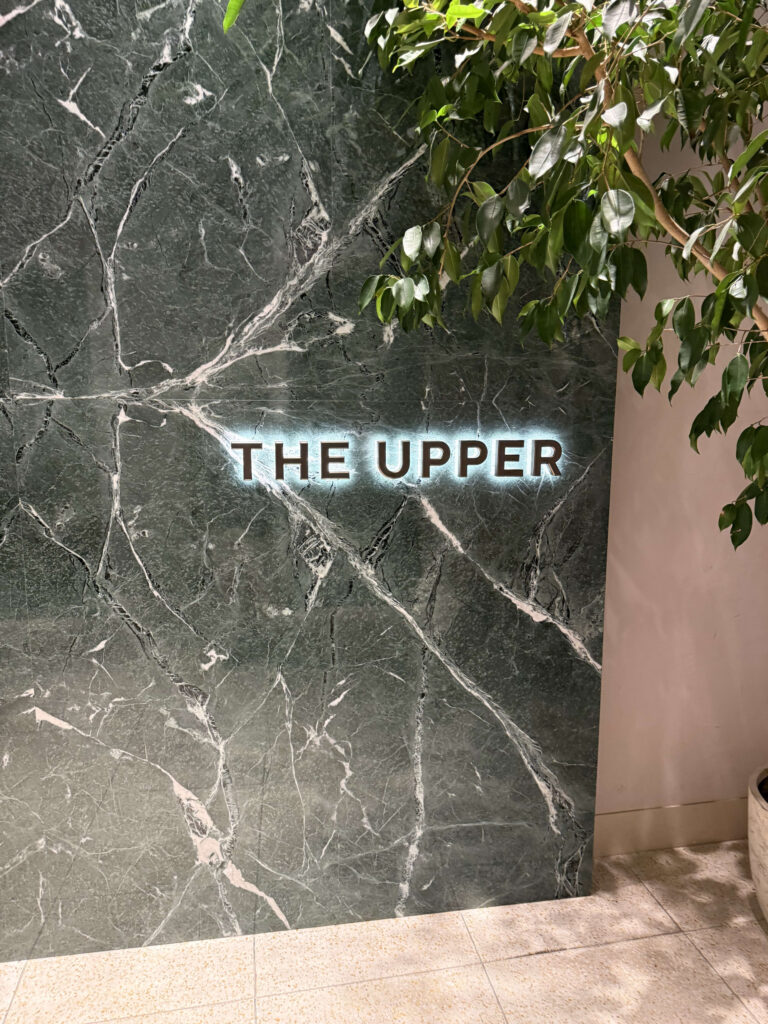 Backlit 'THE UPPER' sign mounted on a dark green marble wall framed by plant leaves