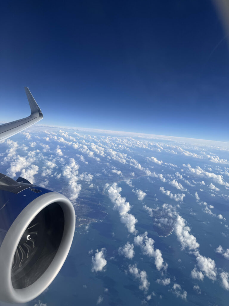 View from an airplane window showing the engine and clouds parting to reveal the ocean and land below