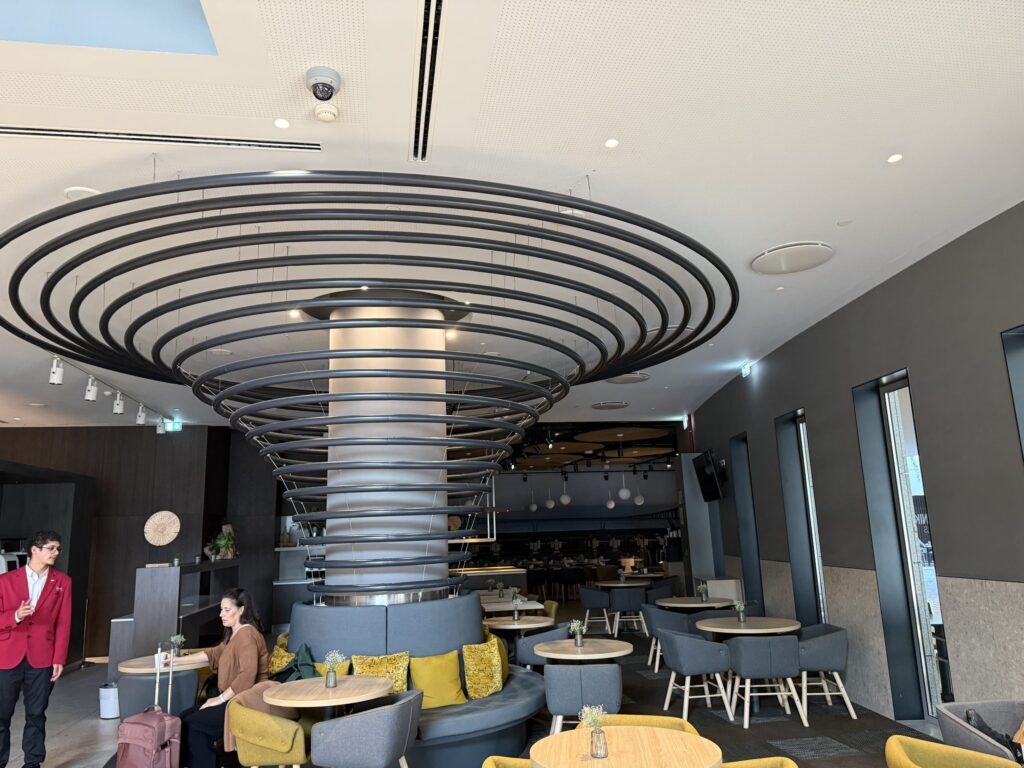 Modern hotel breakfast venue featuring a massive black spiral ceiling sculpture, grey sofas with yellow pillows, and a staff member in a red suit.