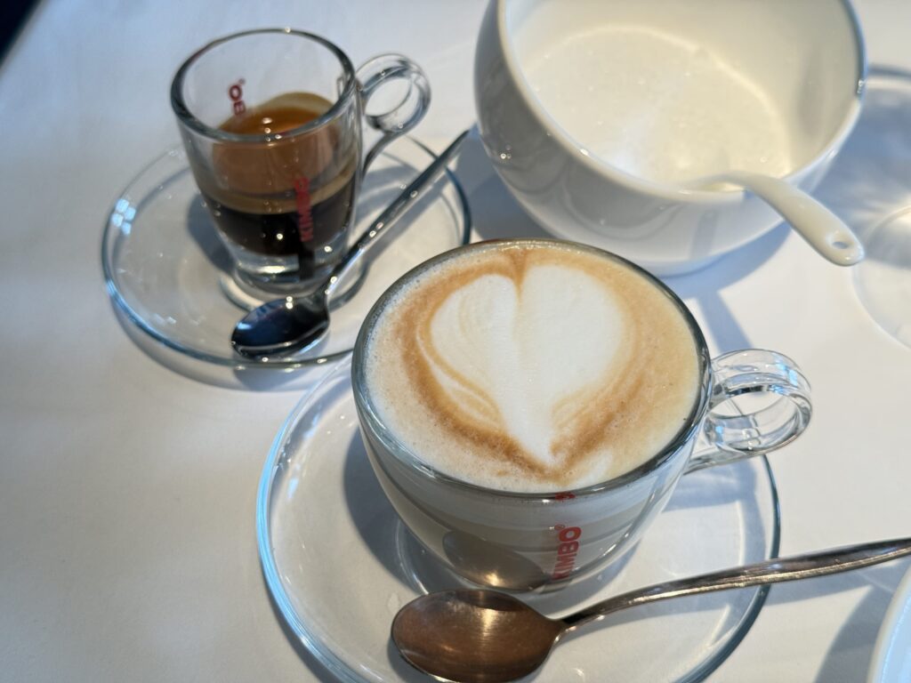A glass cup of cappuccino with heart-shaped latte art, accompanied by an espresso and a sugar bowl in the background