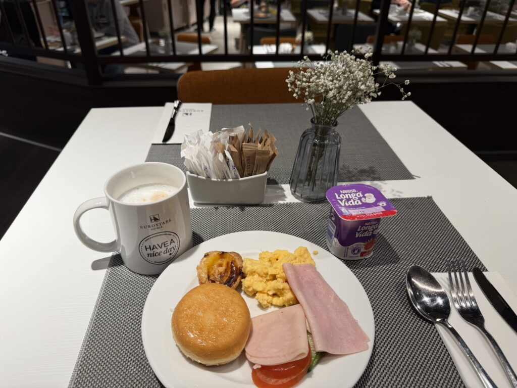 Breakfast table at Eurostars Hotel with a plate of eggs and ham, a coffee mug, yogurt, and a small vase of flowers.