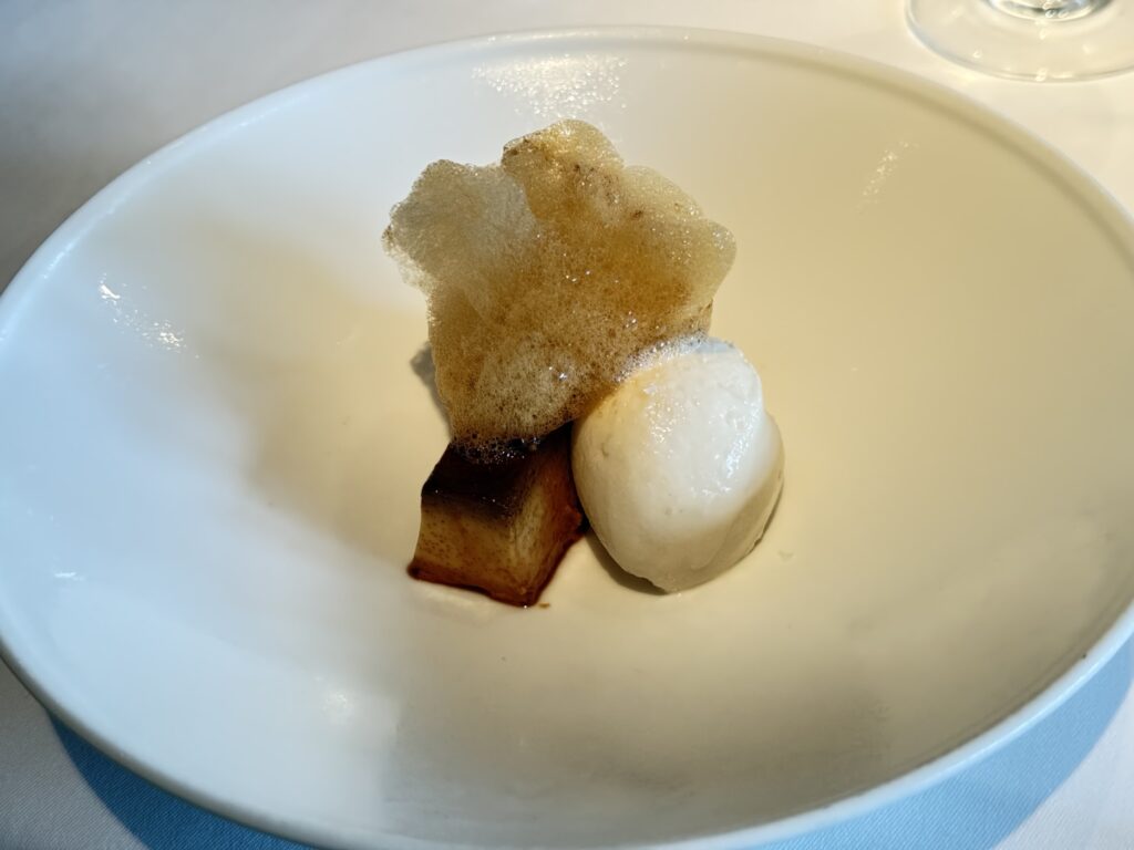 A square piece of homemade pudding and a scoop of salt milk gelato in a white bowl, topped with airy caramel foam