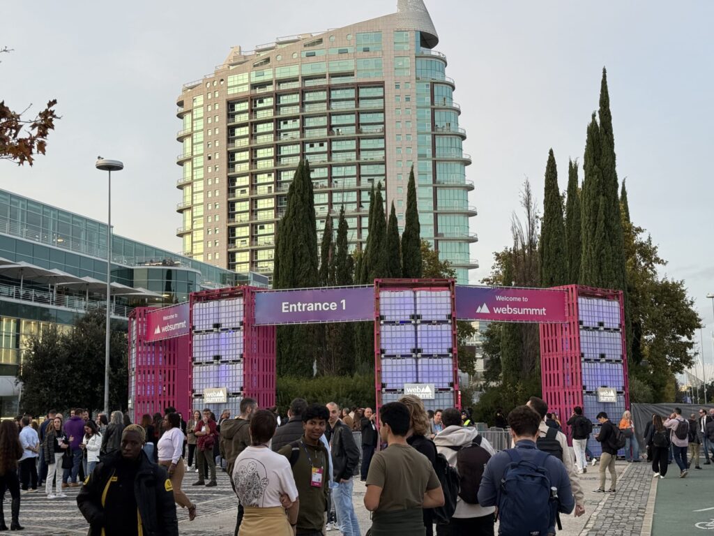 Crowd of people entering the Web Summit via Entrance 1 gate, with a tall glass building in the background.