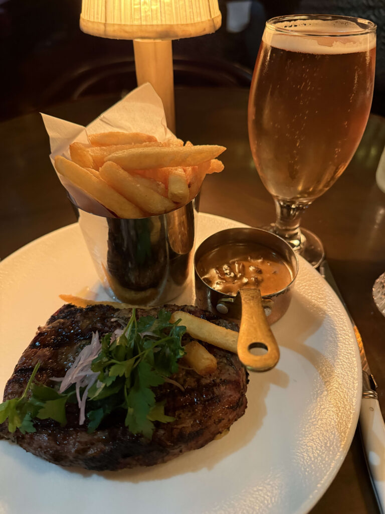 30-day aged ribeye steak on a white plate with french fries in a silver cup, sauce, and a glass of beer
