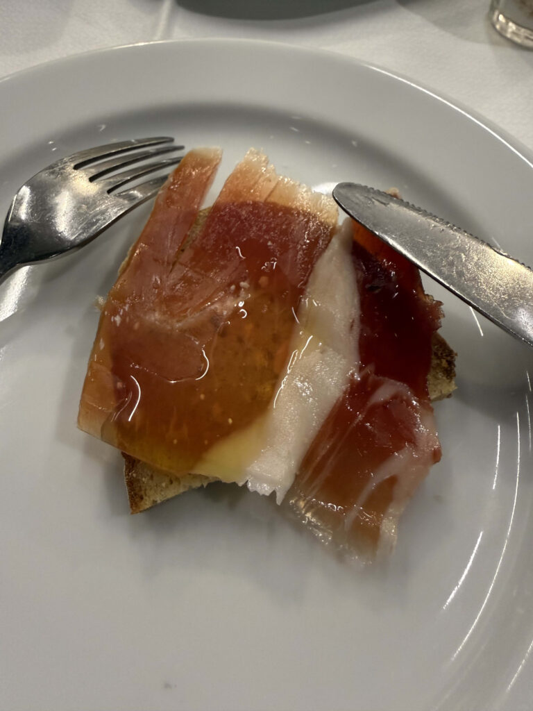 Toast topped with slices of cured ham and olive oil on a white plate with a fork and knife.