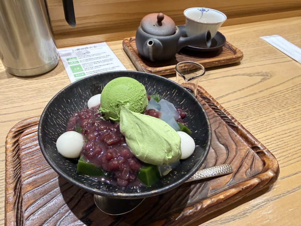 Matcha Cream Anmitsu dessert featuring matcha ice cream, red bean paste, mochi balls, and jelly cubes in a black bowl, served with a tea set on a wooden table.