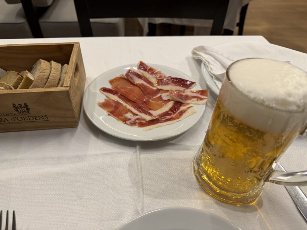 白いテーブルクロスの上に置かれた生ハムの皿、パンが入った木箱、泡の立ったビールジョッキ