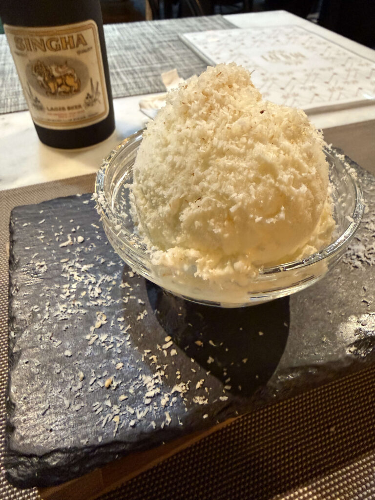 黒いスレートプレートに乗った、ココナッツフレークたっぷりのココナッツアイスクリームと背景のシンハービール
