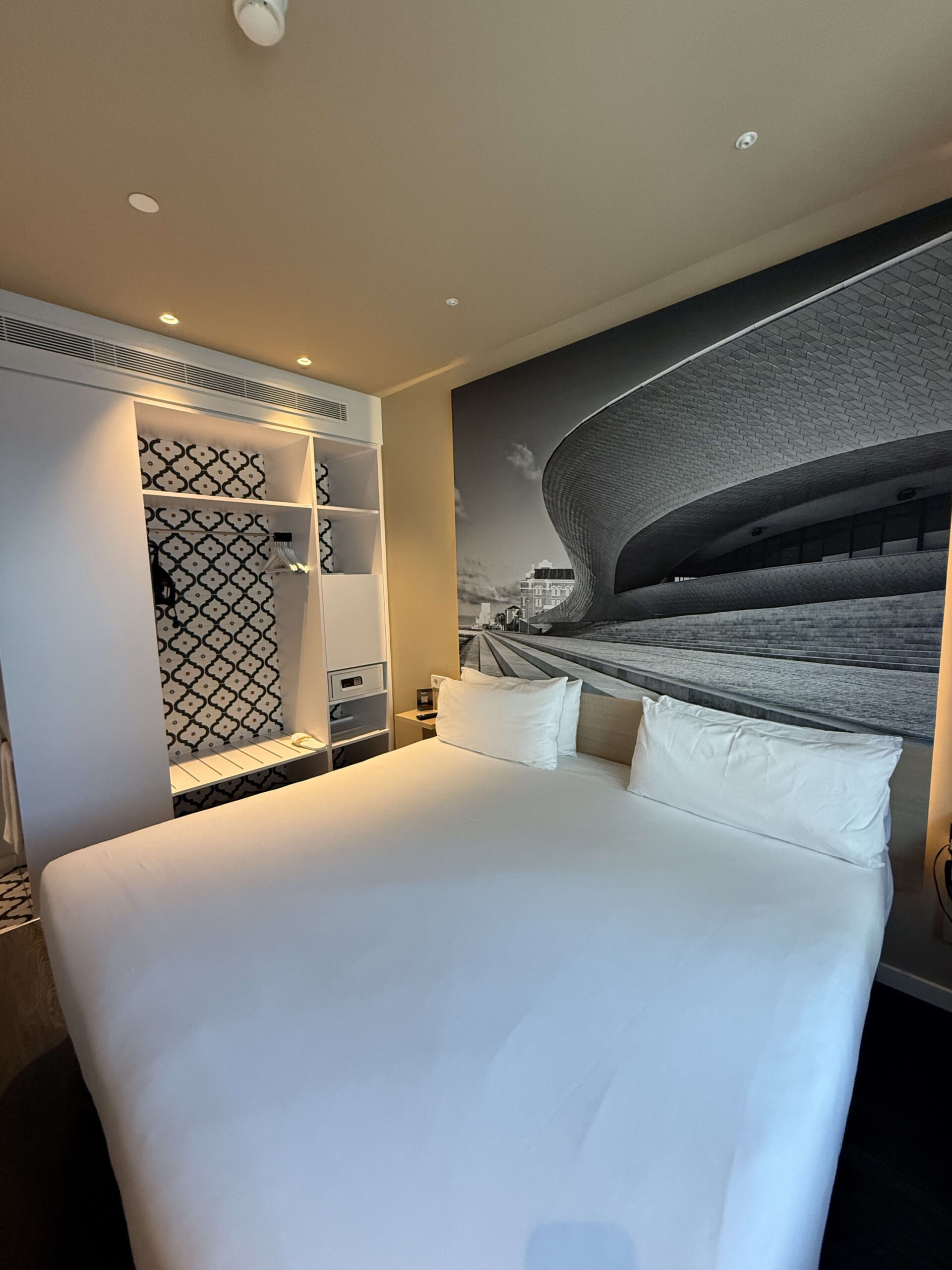 Modern hotel room featuring a white double bed, architectural wall mural, and an open closet with geometric patterns