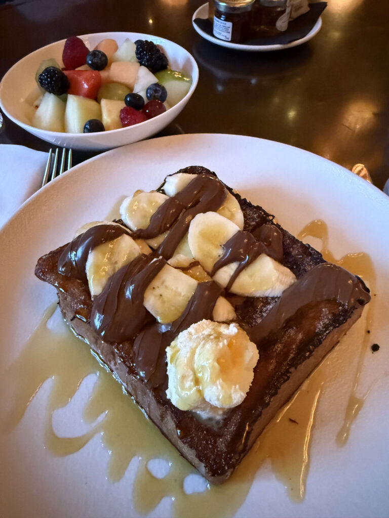 Thick slice of French toast topped with sliced bananas and drizzled with Nutella, accompanied by a bowl of fresh mixed fruit salad.