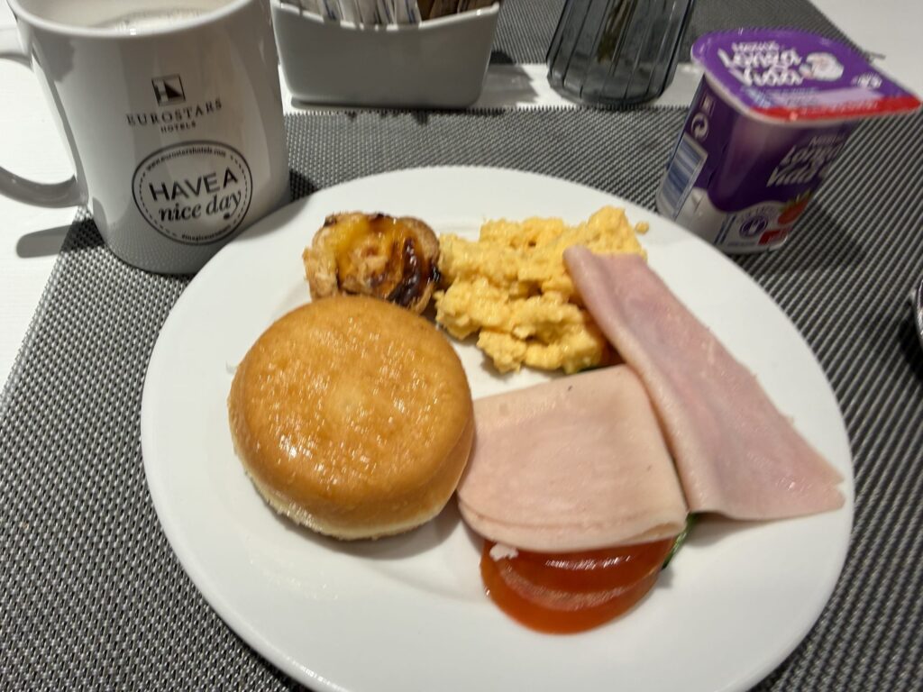 Breakfast plate with donut, egg tart, ham, and scrambled eggs next to a Eurostars Hotels mug