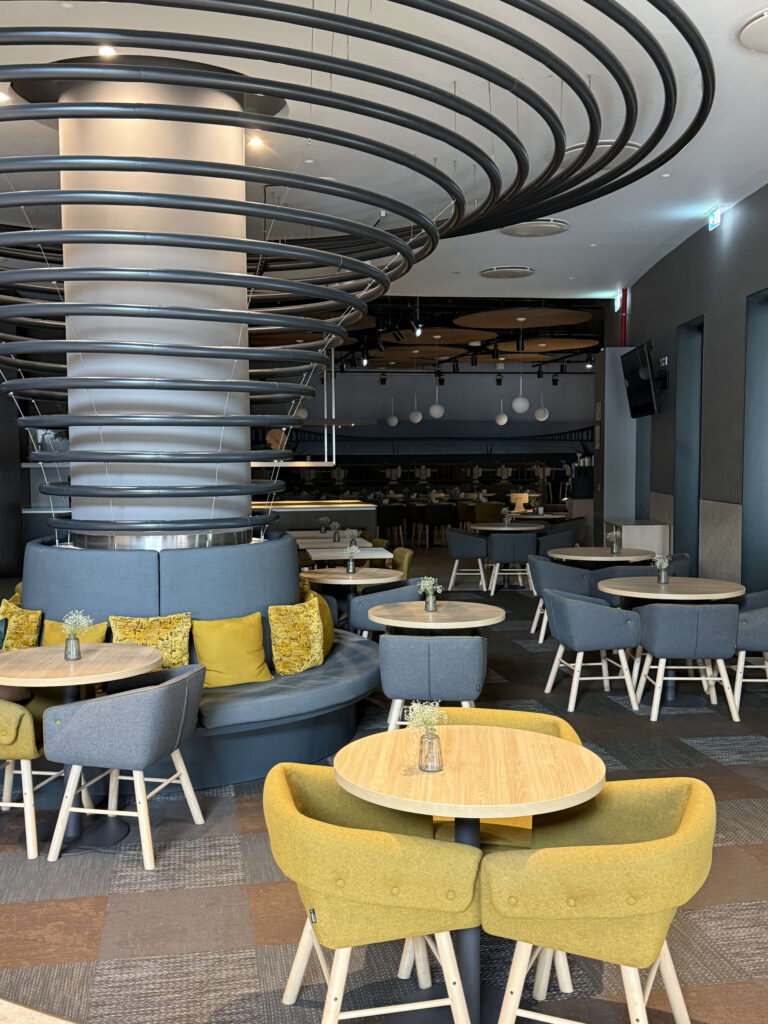 Modern lounge interior featuring a massive spiral ceiling installation, yellow armchairs, grey seating, and round wooden tables.