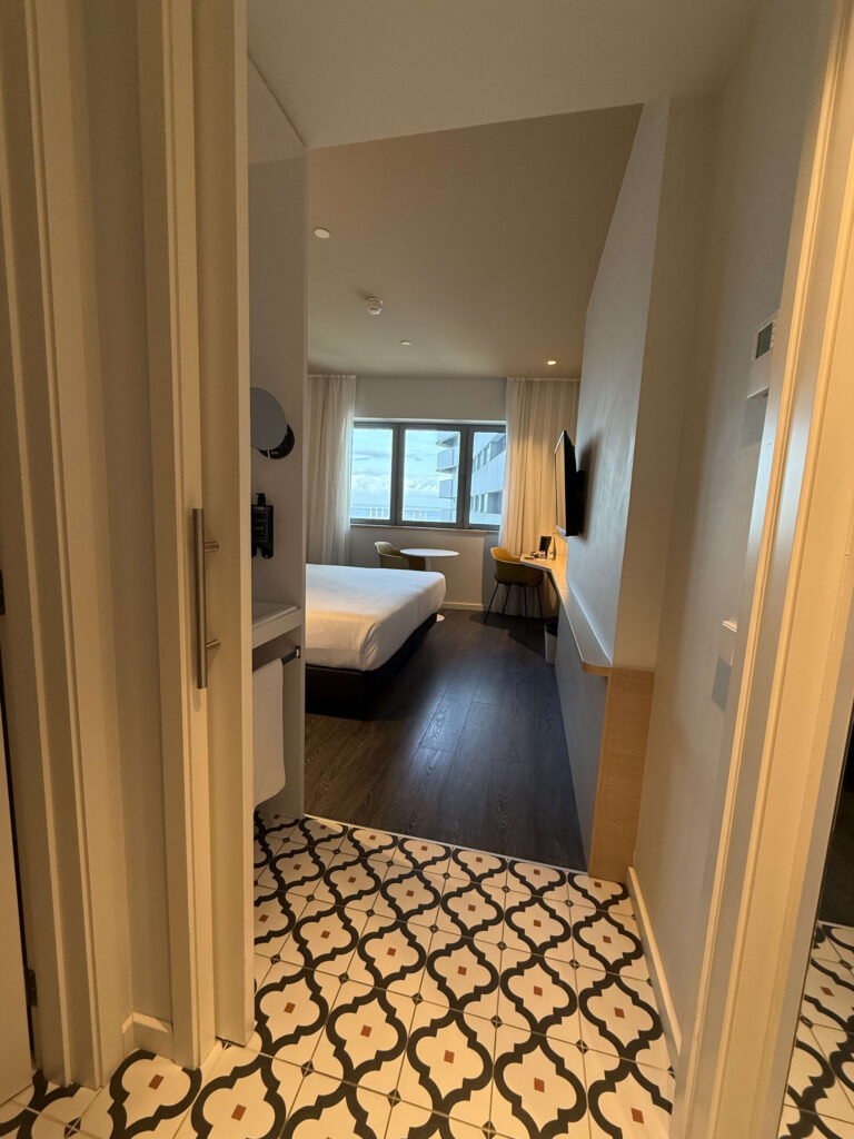 入り口から見たホテルの客室の様子。幾何学模様のタイル床と奥のベッド、窓が見える