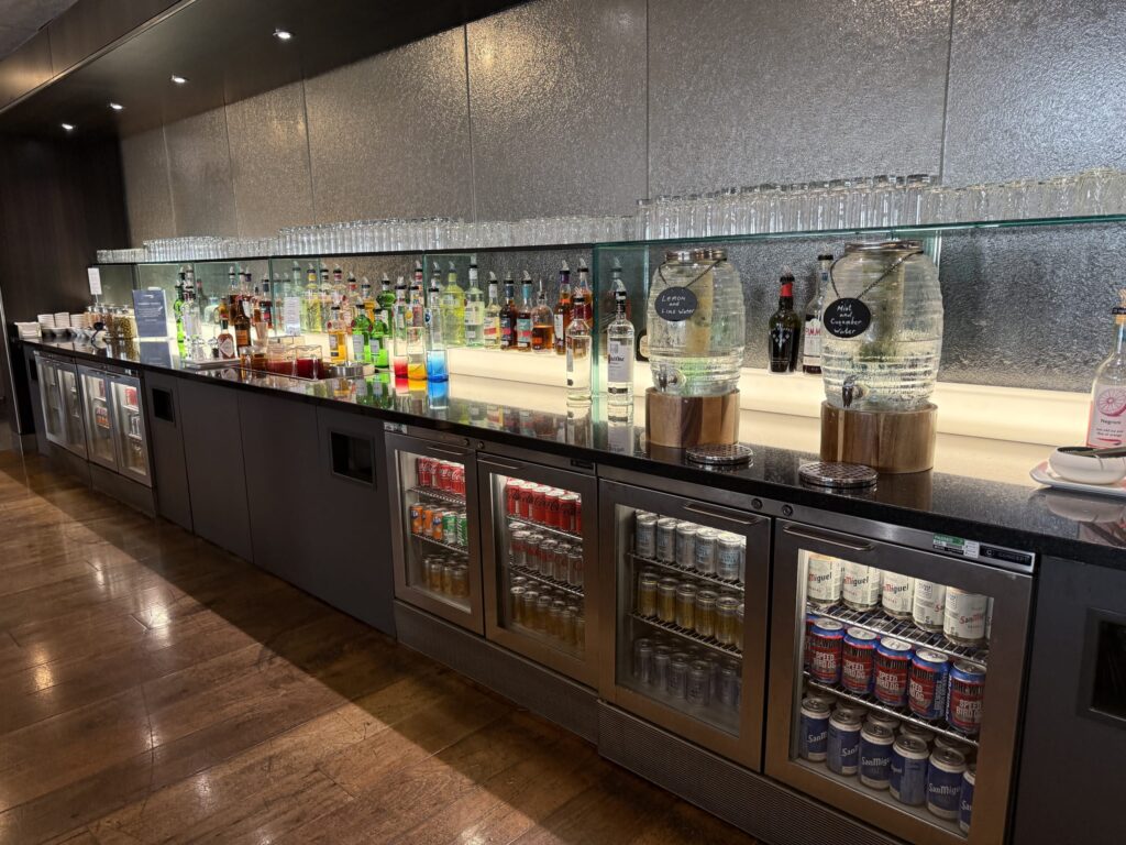 Self-service bar counter in a lounge featuring liquor bottles, flavored water dispensers, and under-counter fridges stocked with beer and soft drinks