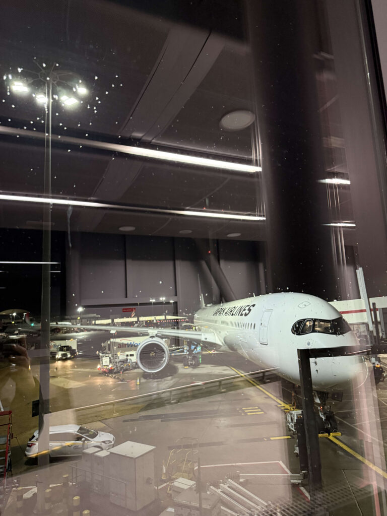 Japan Airlines Airbus A350-1000 parked at a gate at Heathrow Airport at night, viewed through a terminal window